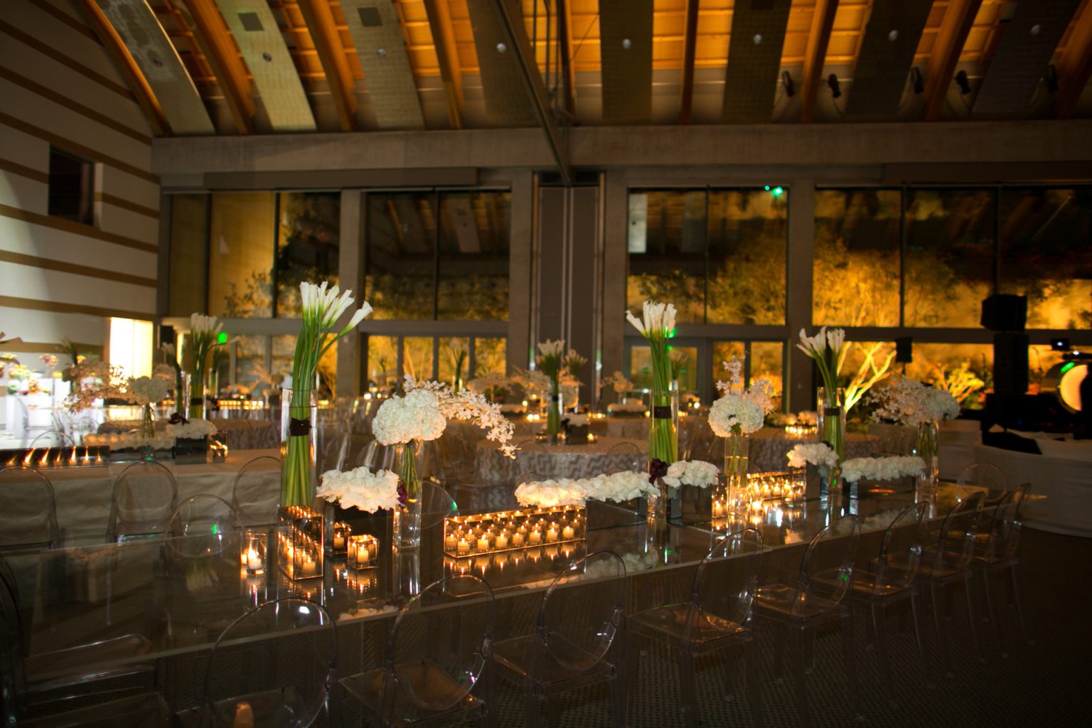 Elegant private event venue with mirrored dinner tables, clear ghost chairs, white floral arrangements, tall calla lilies, and abundant candlelight.