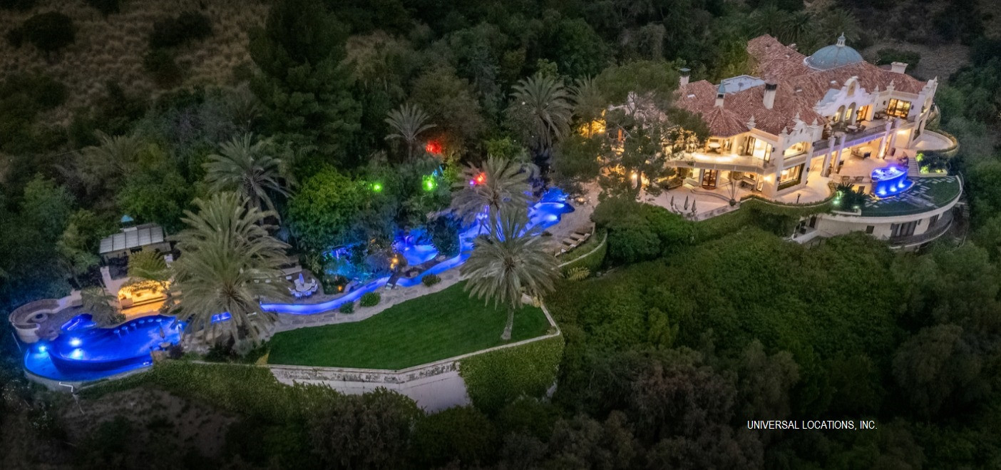 Twilight aerial view of a luxury Mediterranean estate featuring a resort-style pool, tropical gardens, and panoramic Los Angeles views.