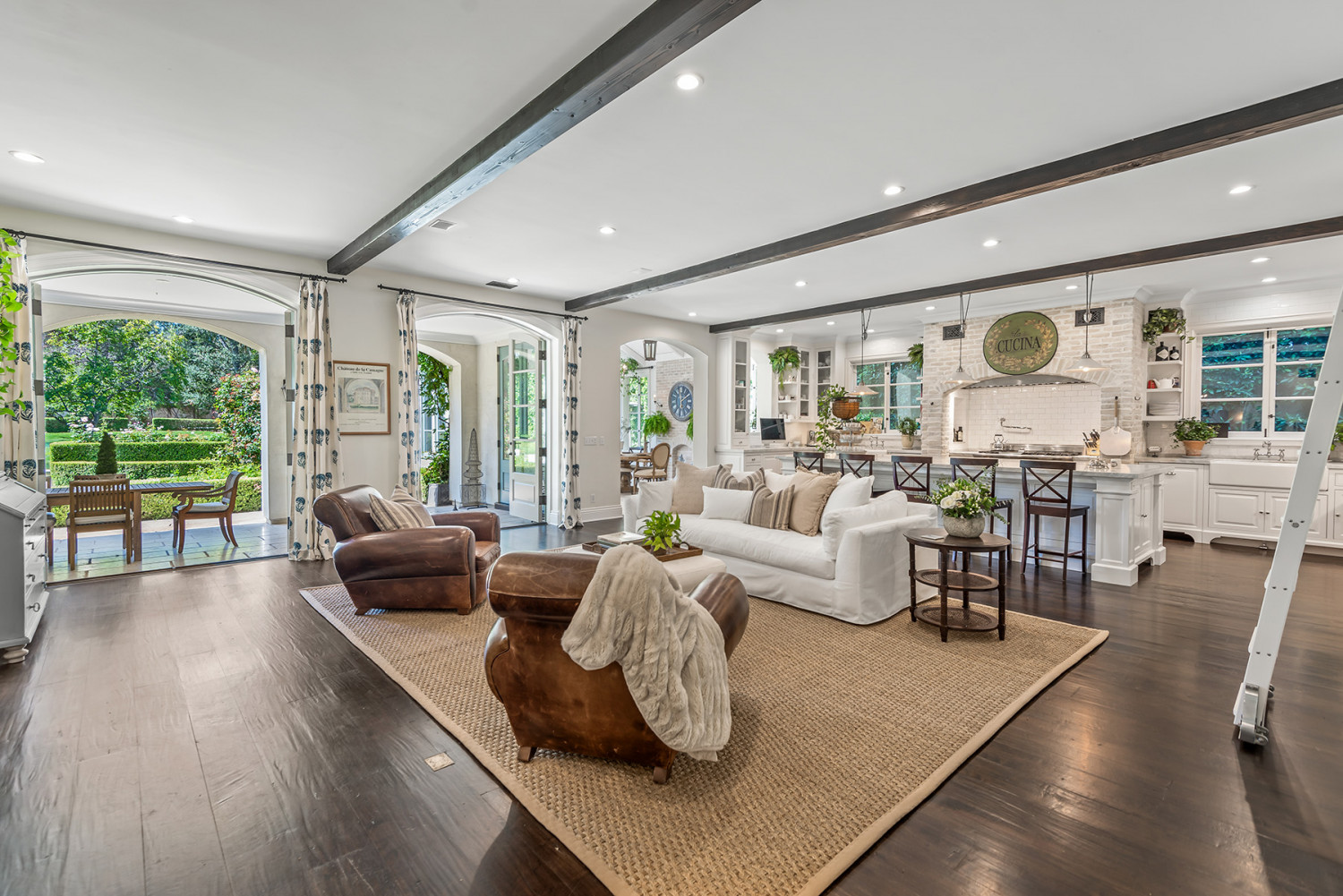 Open living area with a white sofa, leather chairs, dark ceiling beams and an adjoining white kitchen, with doors opening to the garden