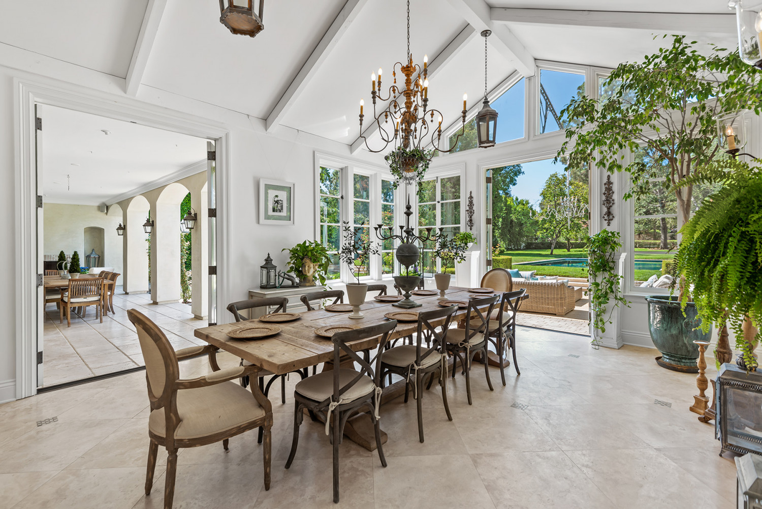 Sunlit dining room with a wooden table, vaulted ceiling, chandelier and tall windows overlooking the pool and garden
