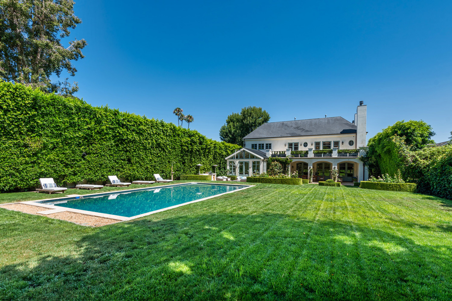 Expansive lawn and rectangular swimming pool framed by tall hedges, with the Encino estate in the background