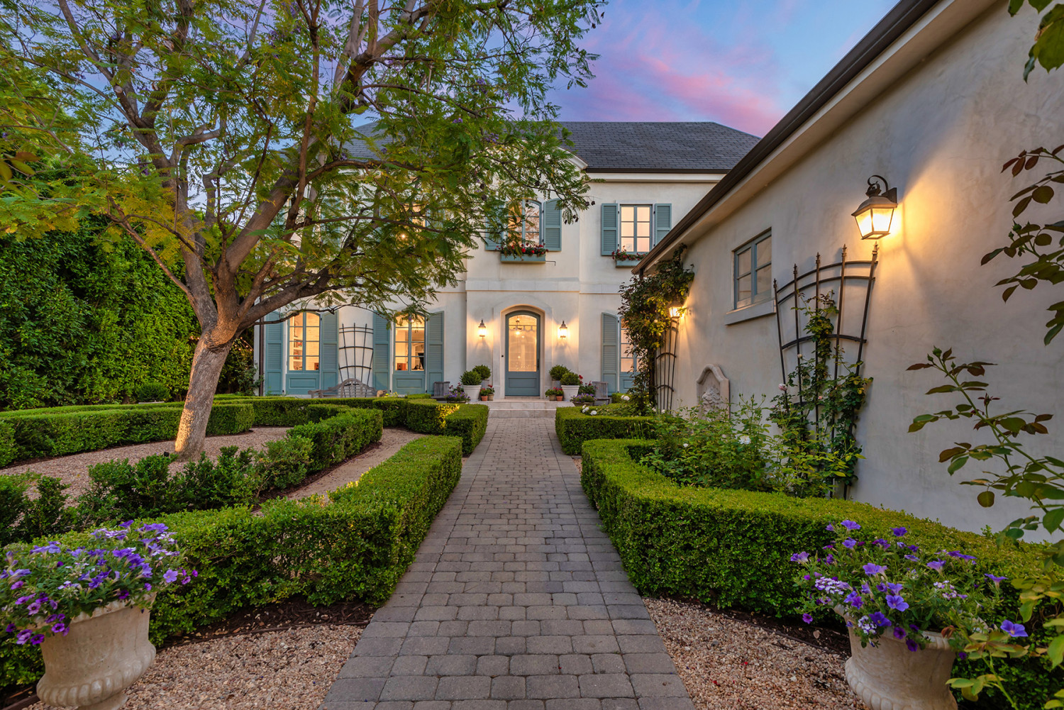 Paved garden walkway bordered by clipped hedges and flowering plants beside the illuminated Encino estate at dusk