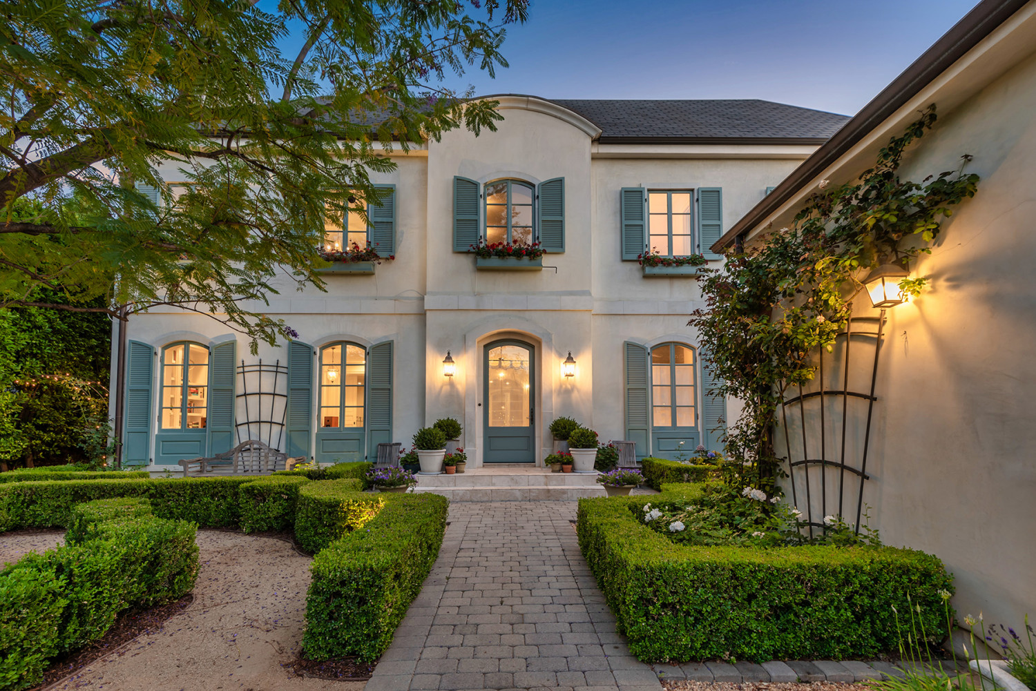 Paved walkway through manicured hedges toward the Encino estate, with illuminated windows and climbing greenery at dusk.