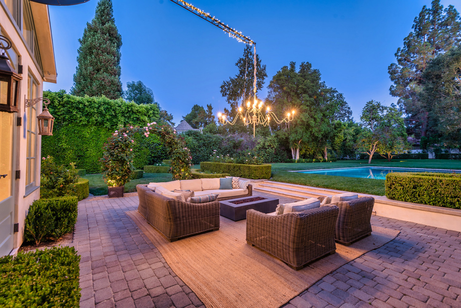 Outdoor lounge seating beneath a suspended chandelier on a paved patio overlooking the pool and tall garden hedges at dusk.