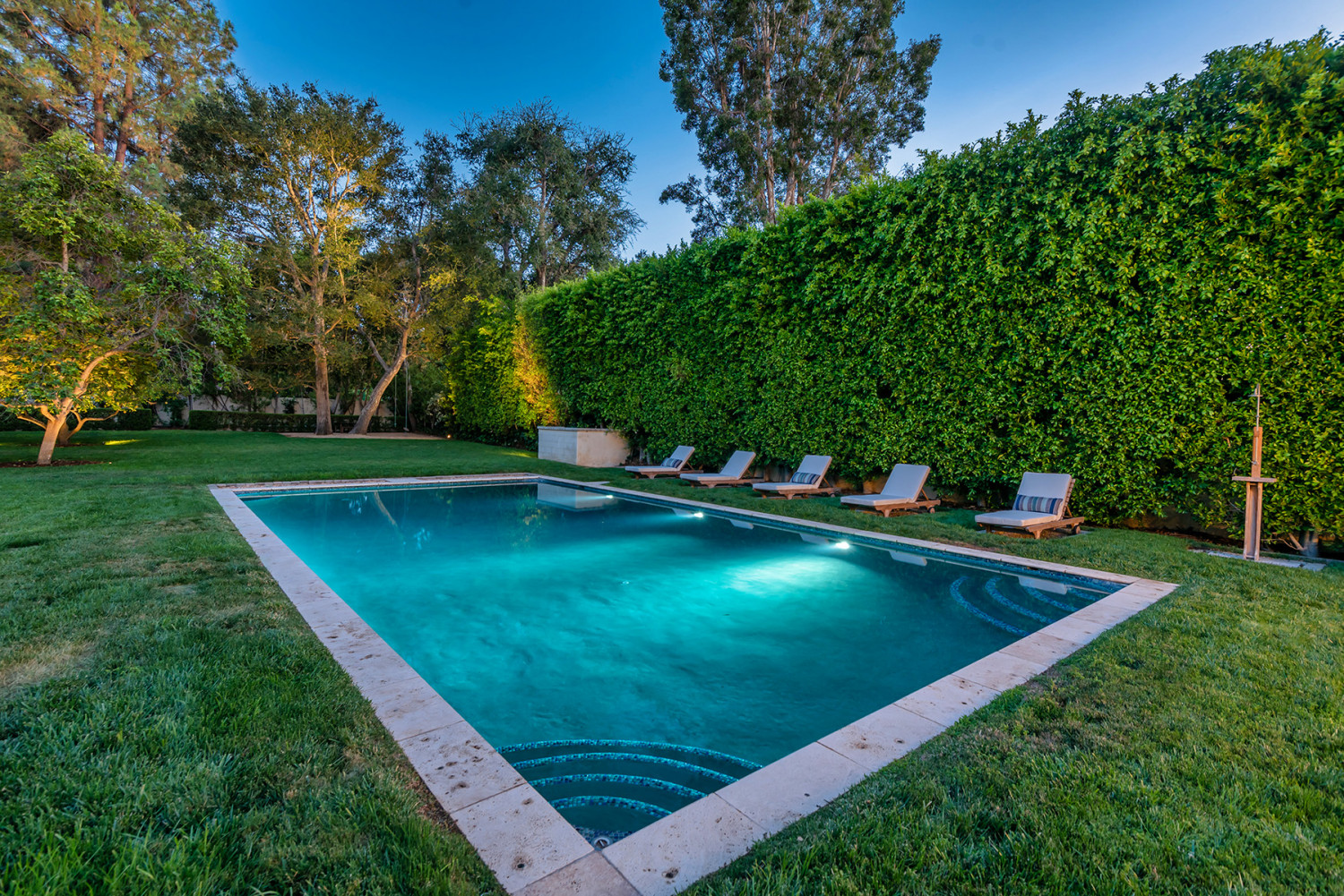 Swimming pool surrounded by lawn, tall hedges and mature trees at dusk
