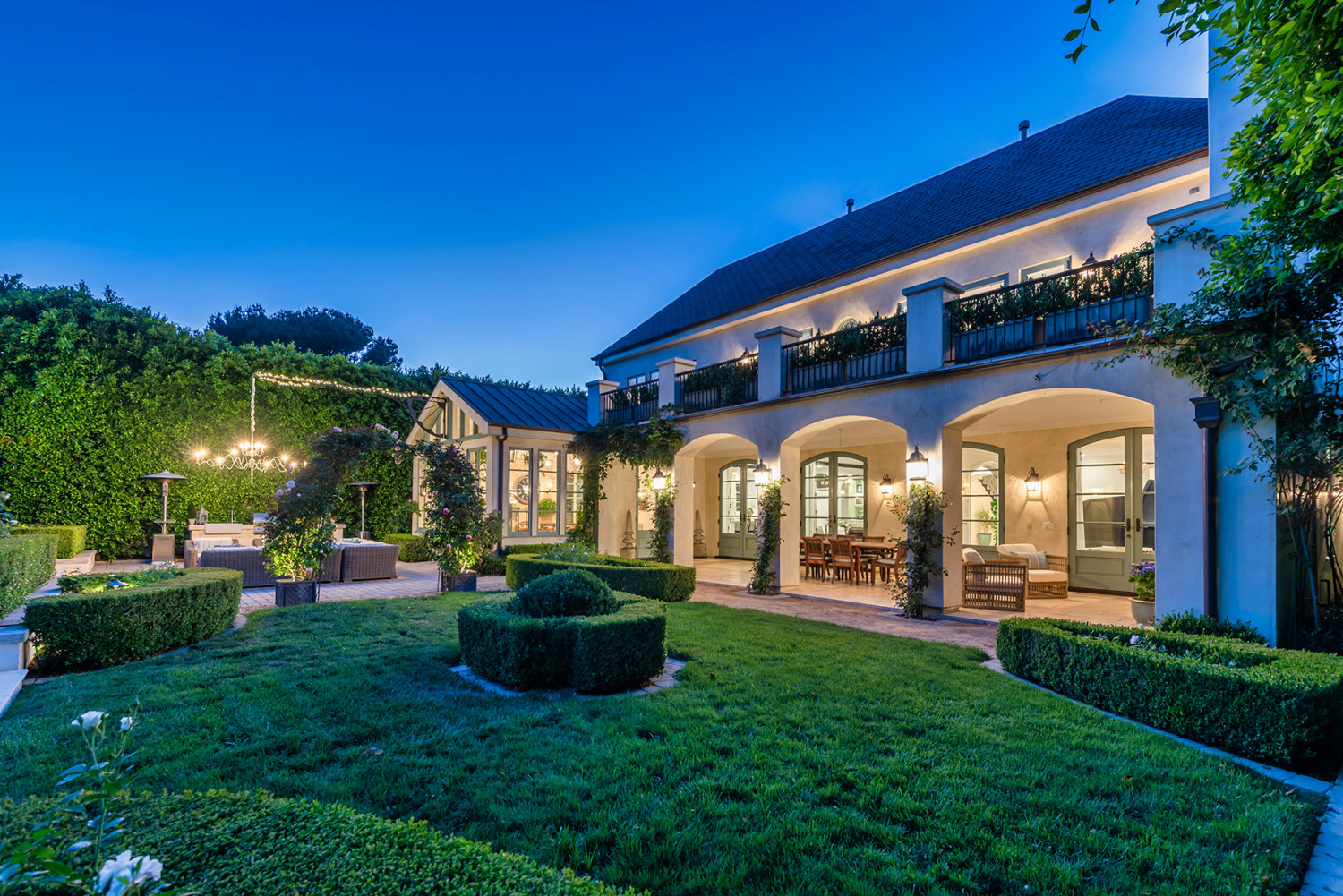 Encino estate at dusk with illuminated arched terraces, manicured garden beds and a lawn.